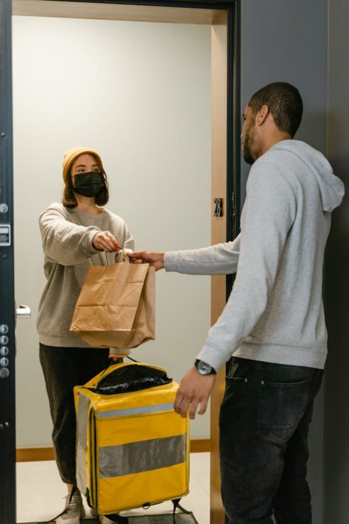 A delivery person hands food to a masked customer at an apartment door, showcasing a contactless exchange.